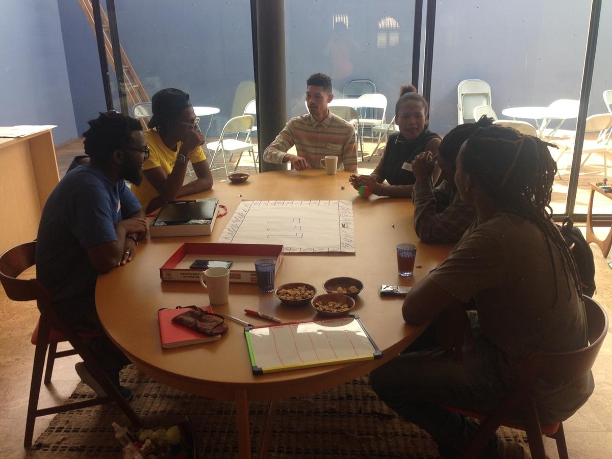 Event photograph from part 2 of Jefferson Bobs Tshabalala’s residency on A4’s top floor. A wooden table hosts a game board drawn out with felt pen markers on a sheet of paper, along with snacks. Six participants sit in discussion around the table.
