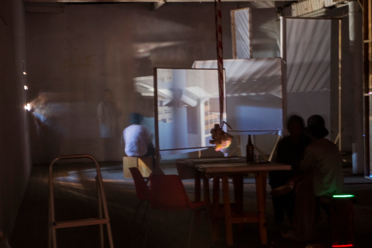 Installation photograph from George Mahashe’s ‘Camera Obscura #3’ exhibition on A4’s top floor. At the back, the darkened space features a freestanding surface with a projected image from A4’s exterior facilitated by Mahashe’s pinhole camera. In the middle, a few people are seated.
