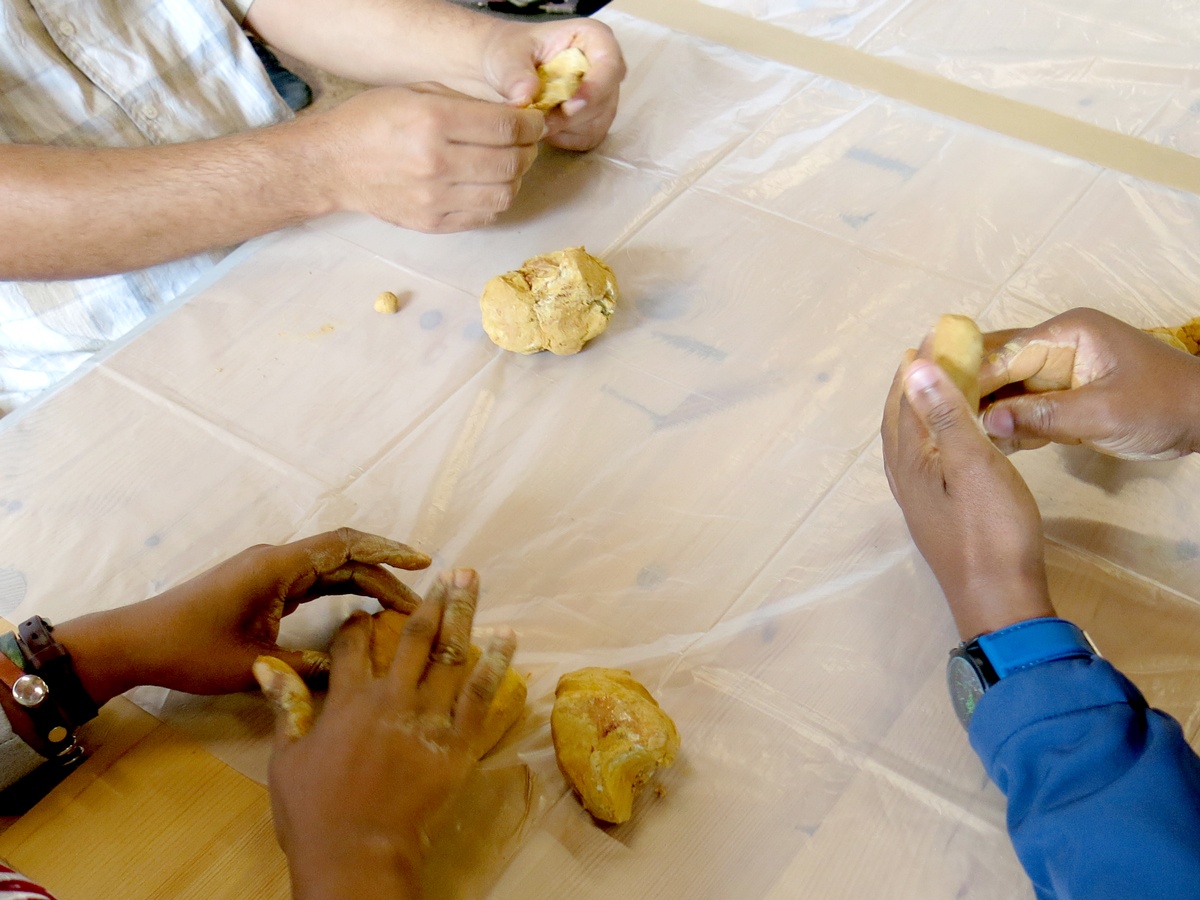 Event photograph from the 2018 rendition of the City Research Studio exchange with the African Centre for Cities. A closeup view of three participants handling clay on a wooden table covered with plastic sheeting.
