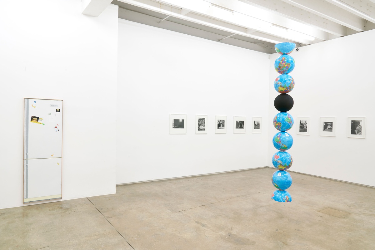 Installation photograph of the Common exhibition. In the middle is Nolan Oswald Dennis’ ‘model for an endless column’, a column of globe models suspended from the ceiling. One is solid black, the first and last are halved. At the back is Sabelo Mlangeni’s ‘Isivumelwano,’ a row of framed black and white photographs intersecting the corner of the room.
