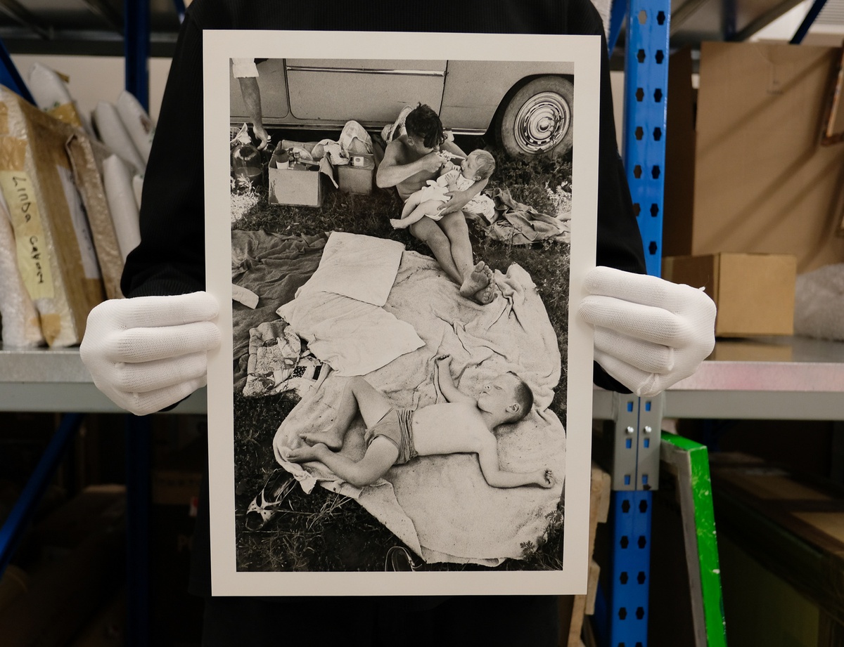 Process photograph from the ‘Picture Theory’ exhibition in A4’s Gallery. Hands wearing archive gloves hold a printed copy of David Goldblatt’s photograph ‘Picnic at Hartebeespoort Dam on New Year's Day, Transvaal (North-West Province) – 3\_B0517’. At the back, a boy holds a toy gun to the head of a baby. At the front, a boy sleeps on a blanket.
