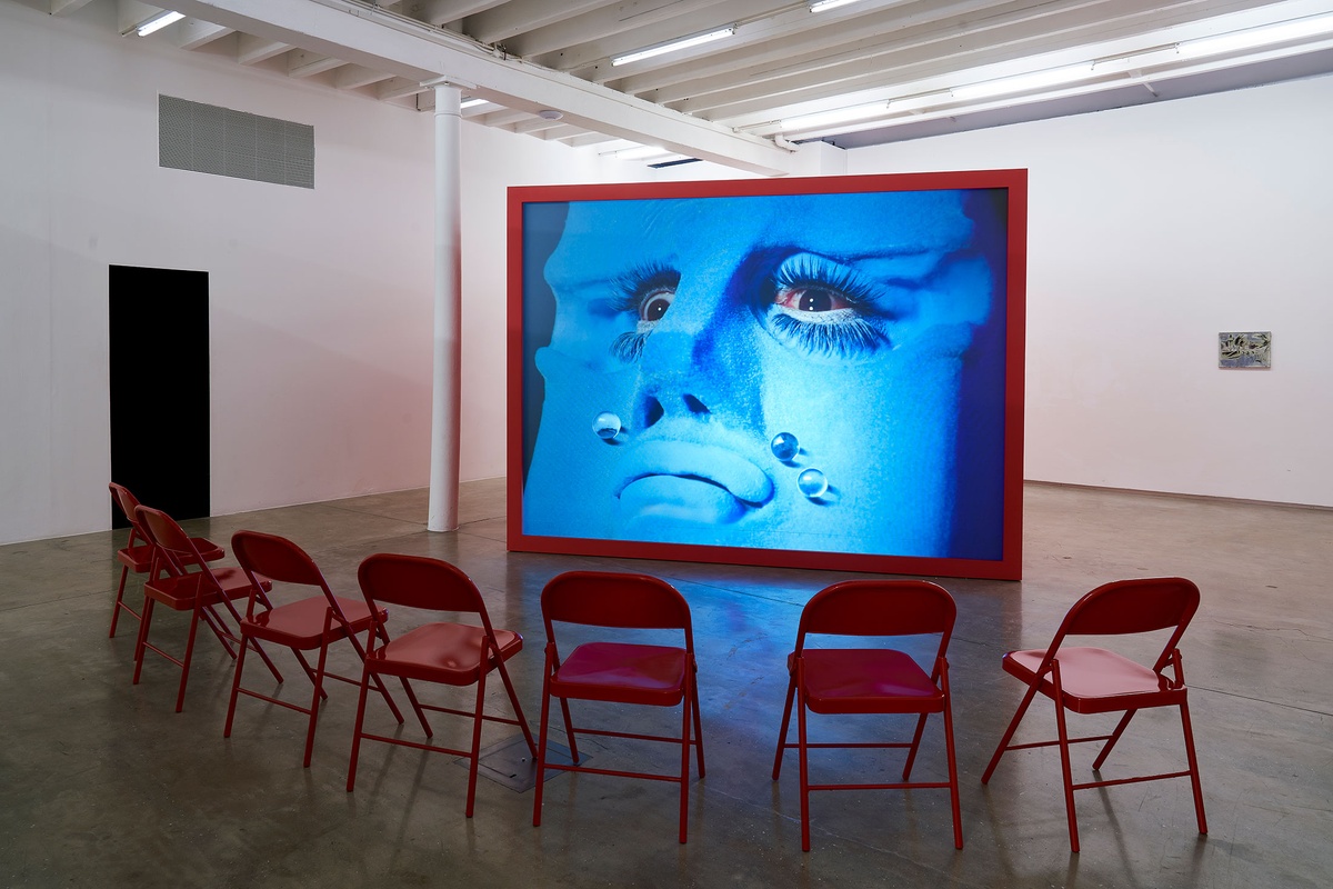 Installation photograph from the 'A Little After This' exhibition in A4 Arts Foundation's gallery that shows Alex Da Corte's video installation 'ROY G BIV'. At the back, a large red wooden box with a back-projected screen plays Da Corte's video. At the front, 7 red powder-coated viewing chairs are arranged in an arch.
