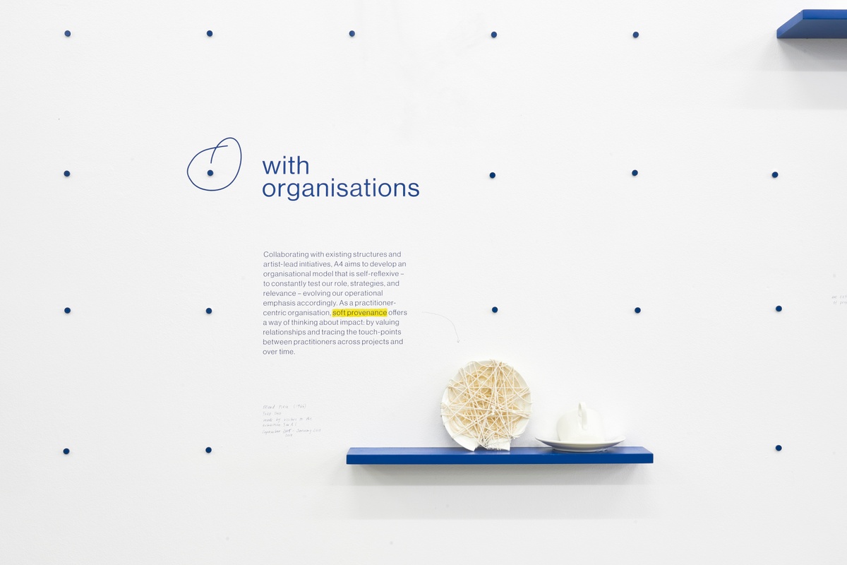 Installation photograph of the A4 About Wall shows the phrase 'with organisations' described and encircled on a white wall in blue. On the left, a short explainer text about A4's approach to collaboration with other organisations. Below, a blue wall-mounted shelf holds sculptural objects produced during Yoko Ono's installation MEND PIECE.
