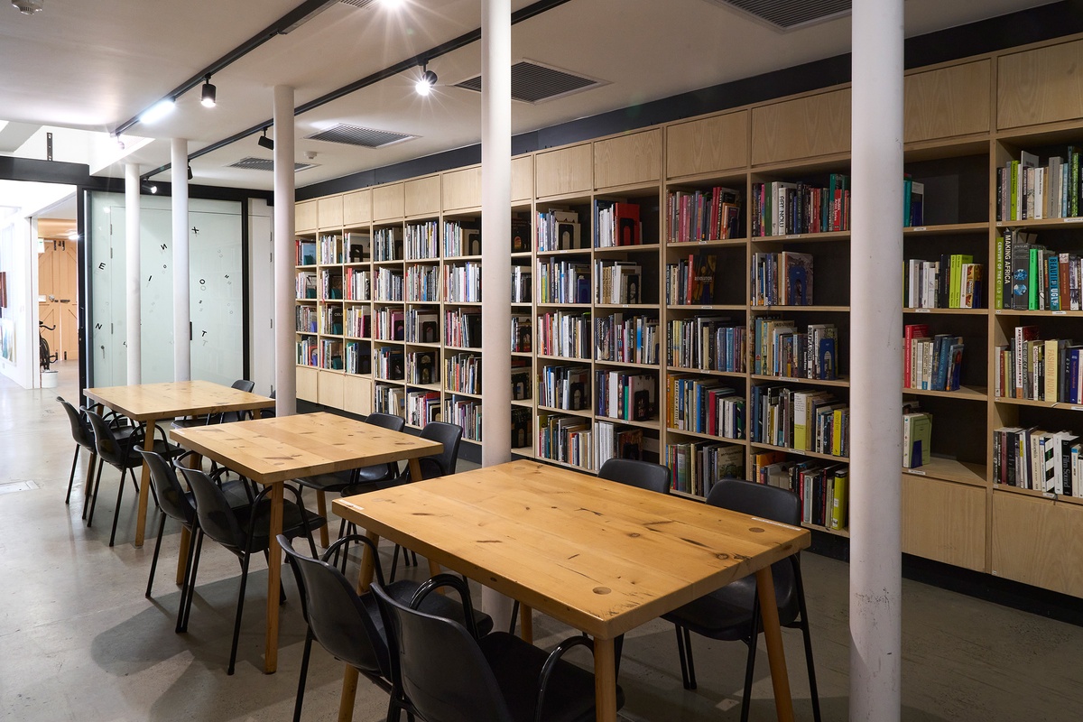 A photograph of the A4 Library. In the middle, a row of rectangular wooden tables with chairs. On the right, a row of book shelves. At the back, a sliding glass door.
