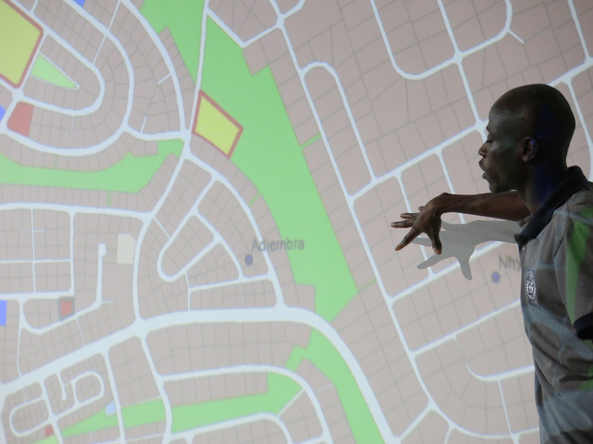 Event photograph from the 2018 rendition of the City Research Studio exchange with the African Centre for Cities. An individual is standing in front of a wall with a projection of a map that indicates the location of Adiembra, Ghana.
