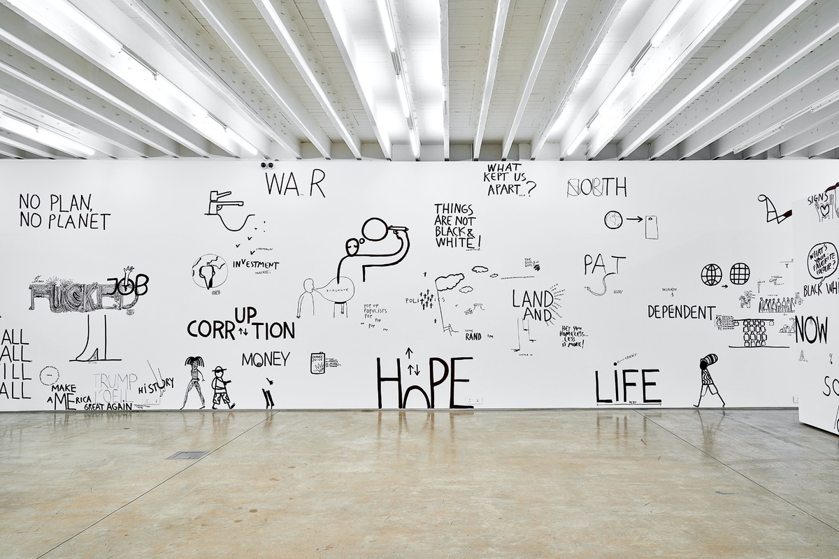 Installation photograph from Dan Perjovschi’s ‘The Black and White Cape Town Report’ exhibition in A4’s Gallery. The white gallery wall is covered in various drawings and phrases in black felt pen marker, including phrases like ‘hope’, ‘life’, ‘war’ and ‘corruption’.
