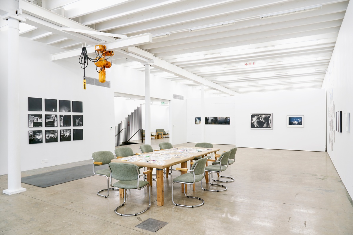 Event photograph from the roundtable discussion that accompanied the ‘Tell It to the Mountains’ exhibition in A4’s gallery. In the middle, a table covered with brown paper and strewn with small printed copies of Lindokuhle Sobekwa and Mikhael Subotzky’s photographs. On the left, Sobekwa’s monochrome photograph ‘Shack of nyaope users is set on fire by an angry group of men that claim they stole their things. Nyaope users are always the first suspects in the communities’ is mounted on the gallery wall. 
