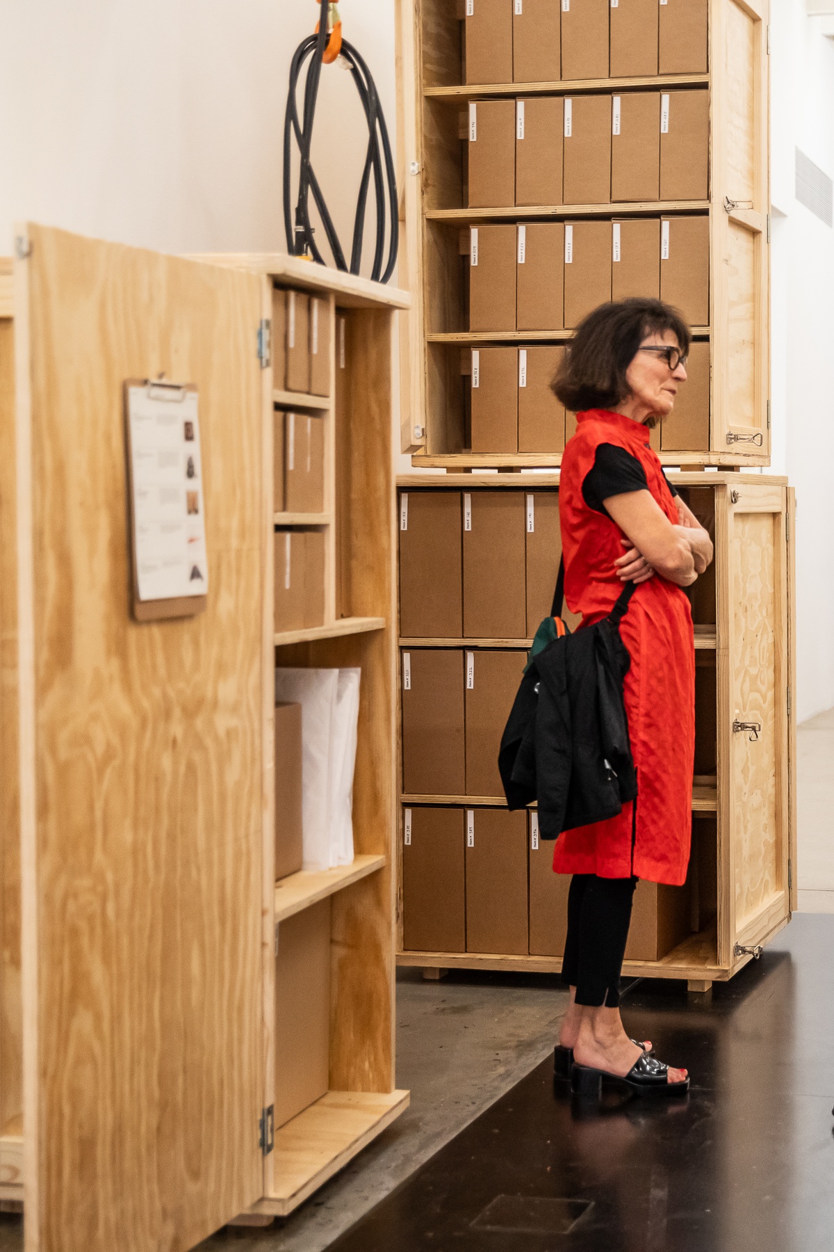 Event photograph from the preview of the 'A Little After This' exhibition in A4 Arts Foundation. At the back, collected objects from Penny Siopis' 'Will' work are stored in envelopes and wooden crates. At the front, Penny Siopis is standing.
