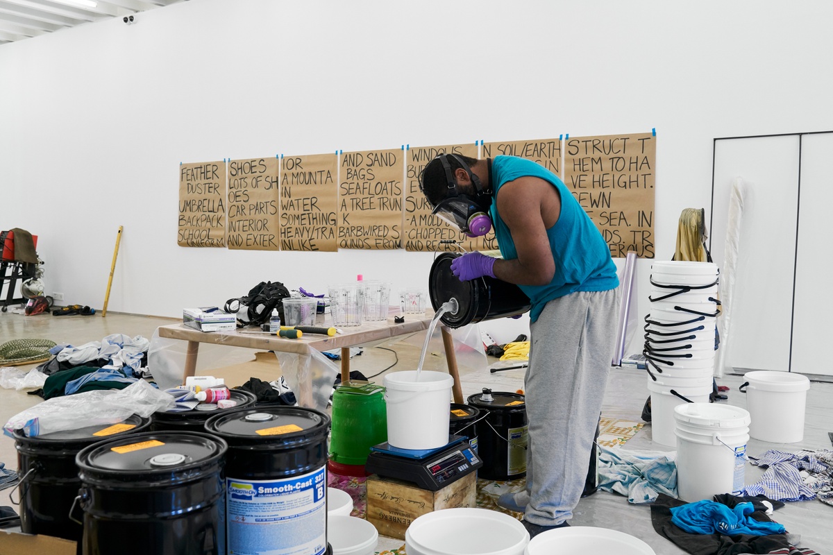 Process photograph from the ‘without a clear discernible image’ exhibition in A4’s Gallery. At the front, Kevin Beasley measures out a liquid component used for the resin hardening process on an electronic scale. At the back, sheets of brown paper with writing are taped to the wall.
