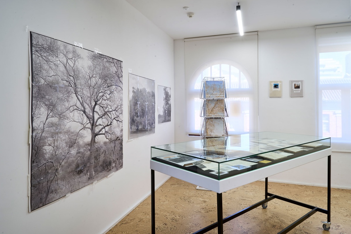 Installation photograph of Michael Tymbios’ studio during his residency in A4 Arts Foundation. At the front, a freestanding display case holds photographs and ephemera. On the left, photographic prints of various sizes are mounted on a white wall. At the back, a freestanding rotating record display stand holds photographic prints.
