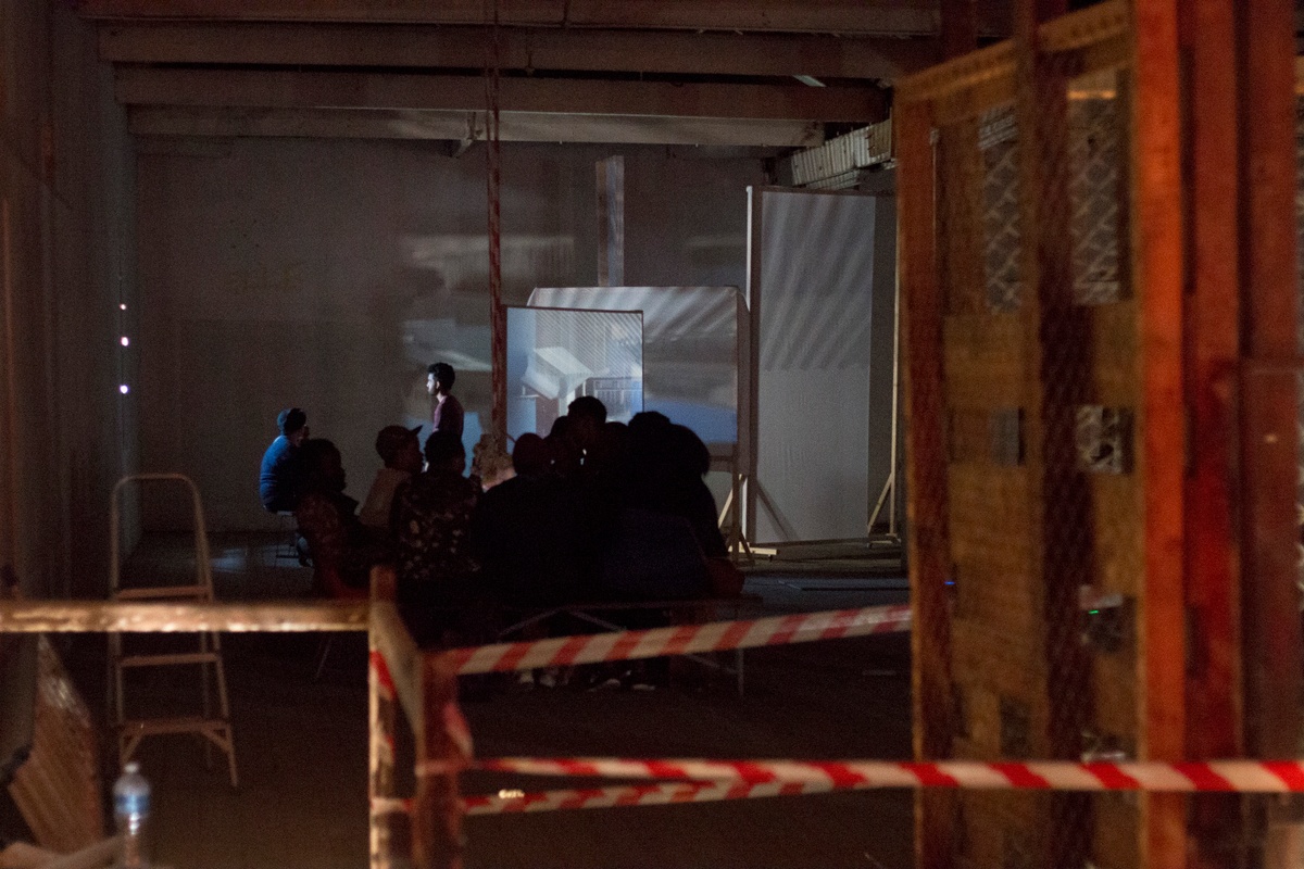 Installation photograph from George Mahashe’s ‘Camera Obscura #3’ exhibition on A4’s top floor. At the back, the darkened space features a freestanding surface with a projected image from A4’s exterior facilitated by Mahashe’s pinhole camera. In the middle, a group of people are seated.
