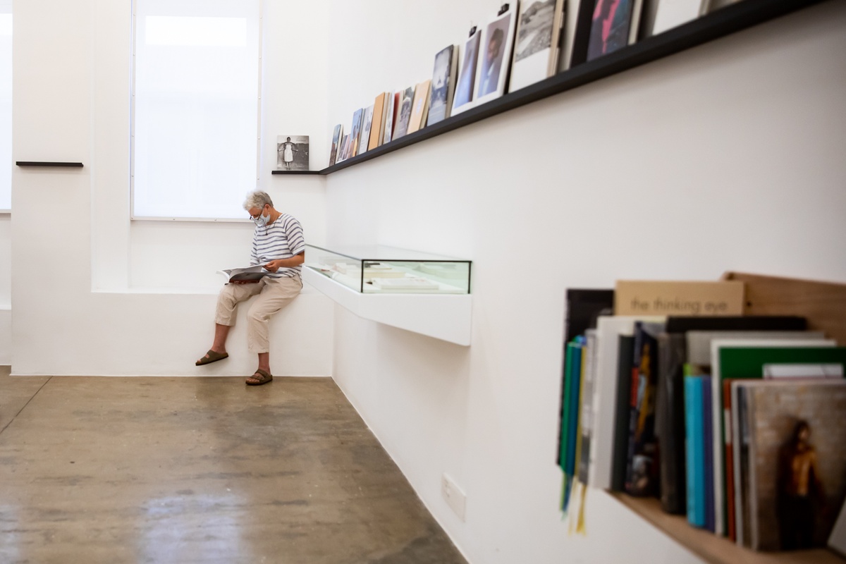Event photograph from the opening of the Photo Book! Photo-Book! Photobook! exhibition in A4’s Gallery in an area dedicated to photobooks from the years 1994 to 2022. In the middle, an attendee sits on a windowsill paging through a book.
