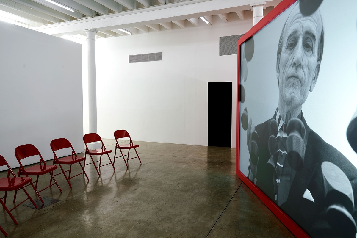 Installation photograph from the 'A Little After This' exhibition in A4 Arts Foundation's gallery that shows Alex Da Corte's video installation 'ROY G BIV'. On the right, a large red wooden box with a back-projected screen plays Da Corte's video. On the left, 7 red powder-coated viewing chairs are arranged in a arch.
