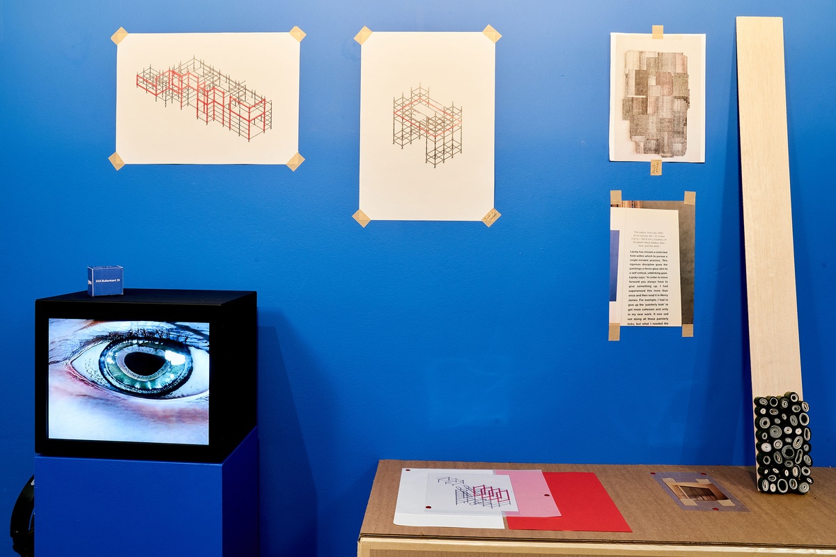 Installation photograph from the ‘mode(l)’ exhibition in A4’s Goods project space. On the left, Nkhensani Mkhari’s video ‘Soft Machine sample’ is displayed on a blue plinth. On the right, a cardboard shelf features Thelma Ndebele’s ‘Groove Bienalle’ prints and Bonolo Kavula’s untitled paper and Shweshwe fabric sculpture.
