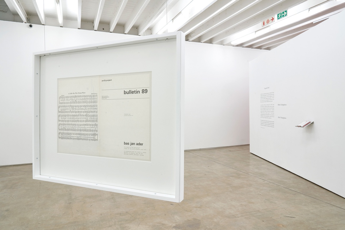 Installation photograph of the You to Me, Me to You exhibition. On the left, Bas Jan Ader’s framed ‘Bulletin 89, in search of the miraculous (songs for the north atlantic)’ is suspended from the ceiling. On the right, Sophie Calle’s ‘Exquisite Pain’ is arranged on a white moveable gallery wall, with a vinyl wall text on the left and a book on a wall-mounted shelf on the right.
