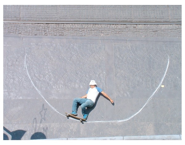 Robin Rhode’s photographic print ‘Catch Air’ depicts a top-down view of stone pavement with a line drawn on it in white to resemble a skateboard ramp. An individual staged with a skateboard appears to be riding the imaginary ramp.
