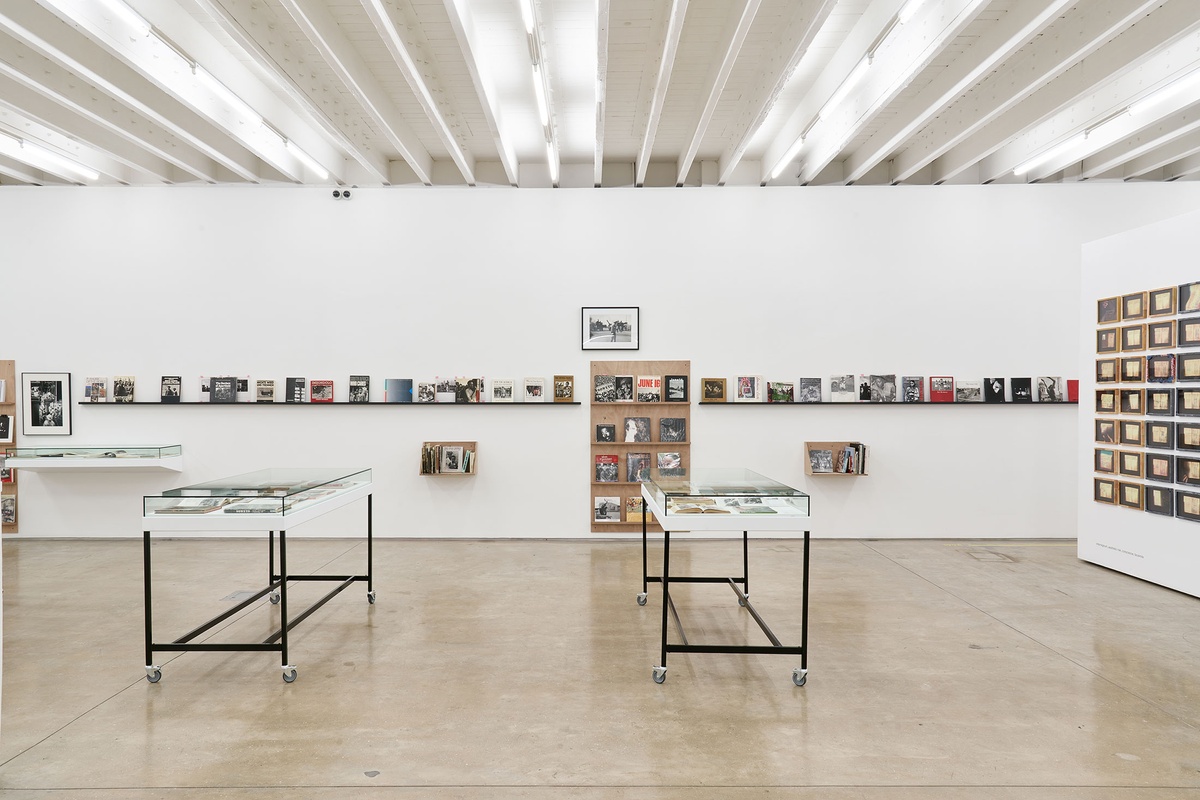 Installation photograph from the Photo Book! Photo-Book! Photobook! exhibition in A4’s Gallery. In the middle, Peter Magubane’s framed photograph ‘Give peace a chance, Soweto’ hangs above a display shelf in an area dedicated to photobooks from the years 1967 to 1994.
