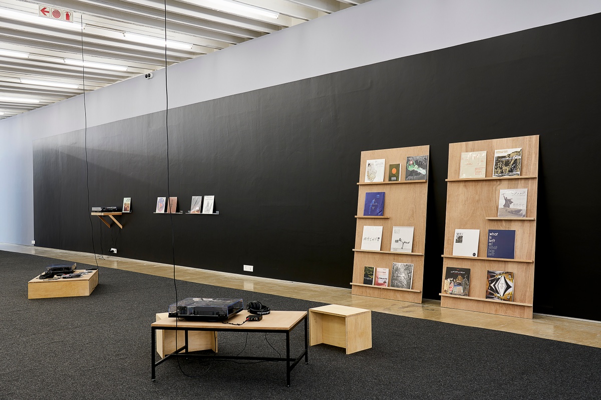 Installation photograph from the ‘Sounding the Void, Imaging the Orchestra V.1’ exhibition in A4’s Gallery. On the left, a collection of vinyls and cassette tapes from the Sahel Sounds label sit on wall mounted shelves. On the right, Bhavisha Panchia’s installation ‘Nothing to Commit Records’ consists of two wooden shelving units with vinyls records that lean against the gallery wall. At the front, two listening areas with record players and earphones sit on the gallery floor.
