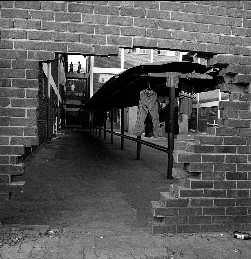 Monochrome photograph ‘Another entrance’ from Sabelo Mlangeni’s residency on A4’s top floor that shows a wall with missing bricks that reveals a building courtyard with a roofed clothesline running along it.
