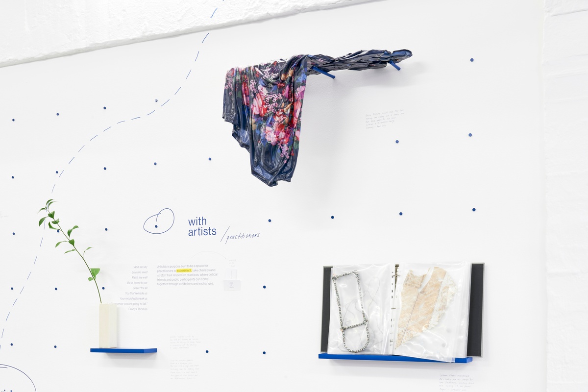 Installation photograph of the A4 About Wall shows the phrase 'with artists/practitioners' described on a white wall in blue. Above, Kevin Beasley's untitled housedress and resin sculpture is mounted on blue dowels. On the right, a folder with process objects by Igshaan Adams. On the left, a branch with leaves in a vase.
