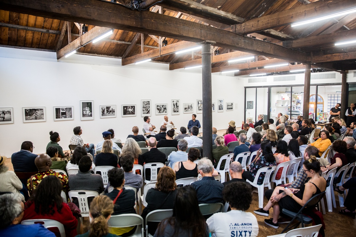 Event photograph from the launch of Omar Badsha’s book ‘Seedtimes’ on A4’s top floor. At the front, audience members are seated in rows. In the middle, the panellists Omar Bash, Imraan Coovadia and Ari Sitas are seated in conversation. At the back, a row of framed monochrome photographs line the white wall behind the panellists.
