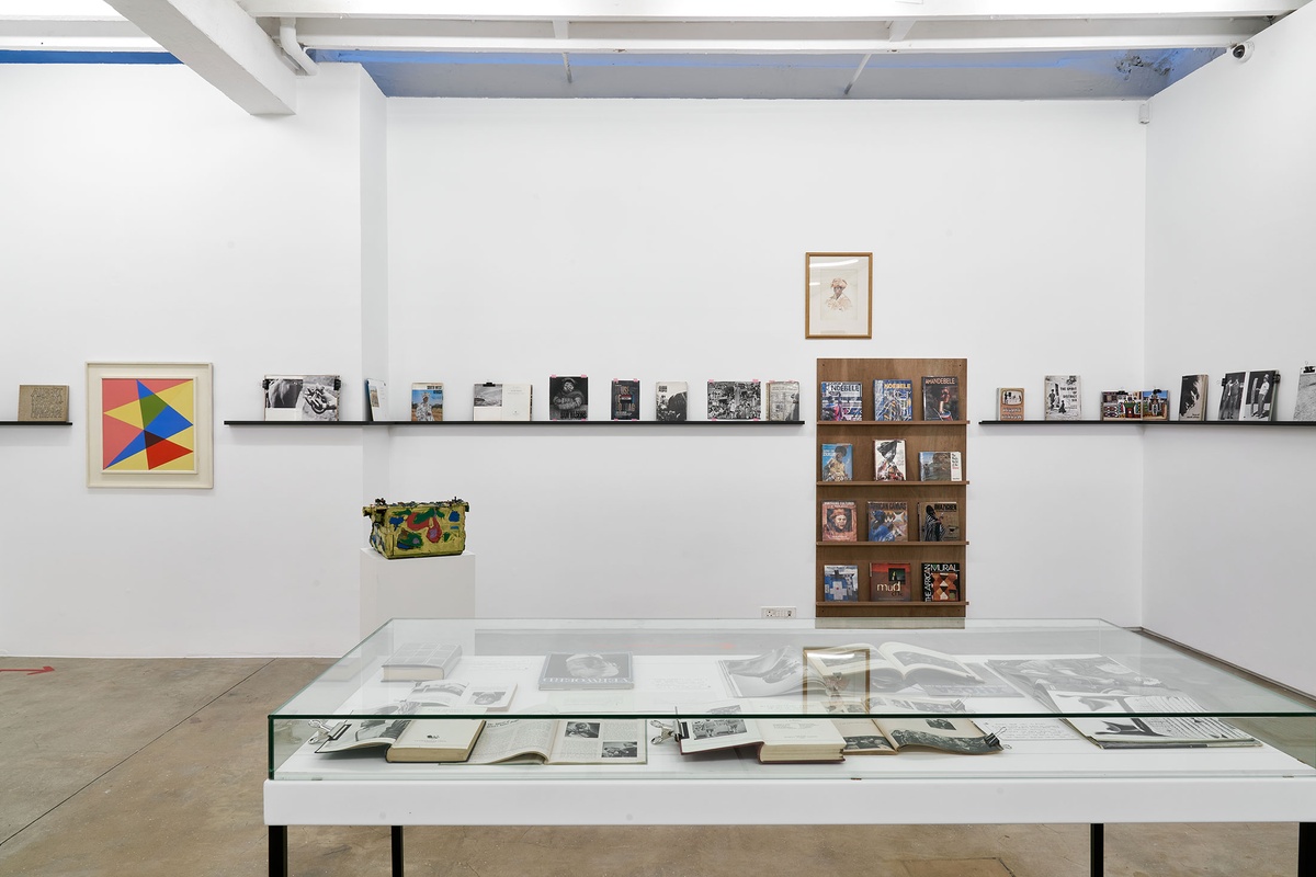 Installation photograph from the Photo Book! Photo-Book! Photobook! exhibition in A4’s Gallery. At the front, a freestanding glass display case with printed matter. In the middle, Walter Battiss’ oil and mixed media work ‘My Typewriter’ sits on a white plinth. At the back, shelving units with printed matter from the years 1967 to 1994 line a white gallery wall, along with Albert Newall’s oil painting ‘Harmonic Development within a Square’ and George Pemba’s watercolour painting ‘Basotho Woman with Headdress’. 
