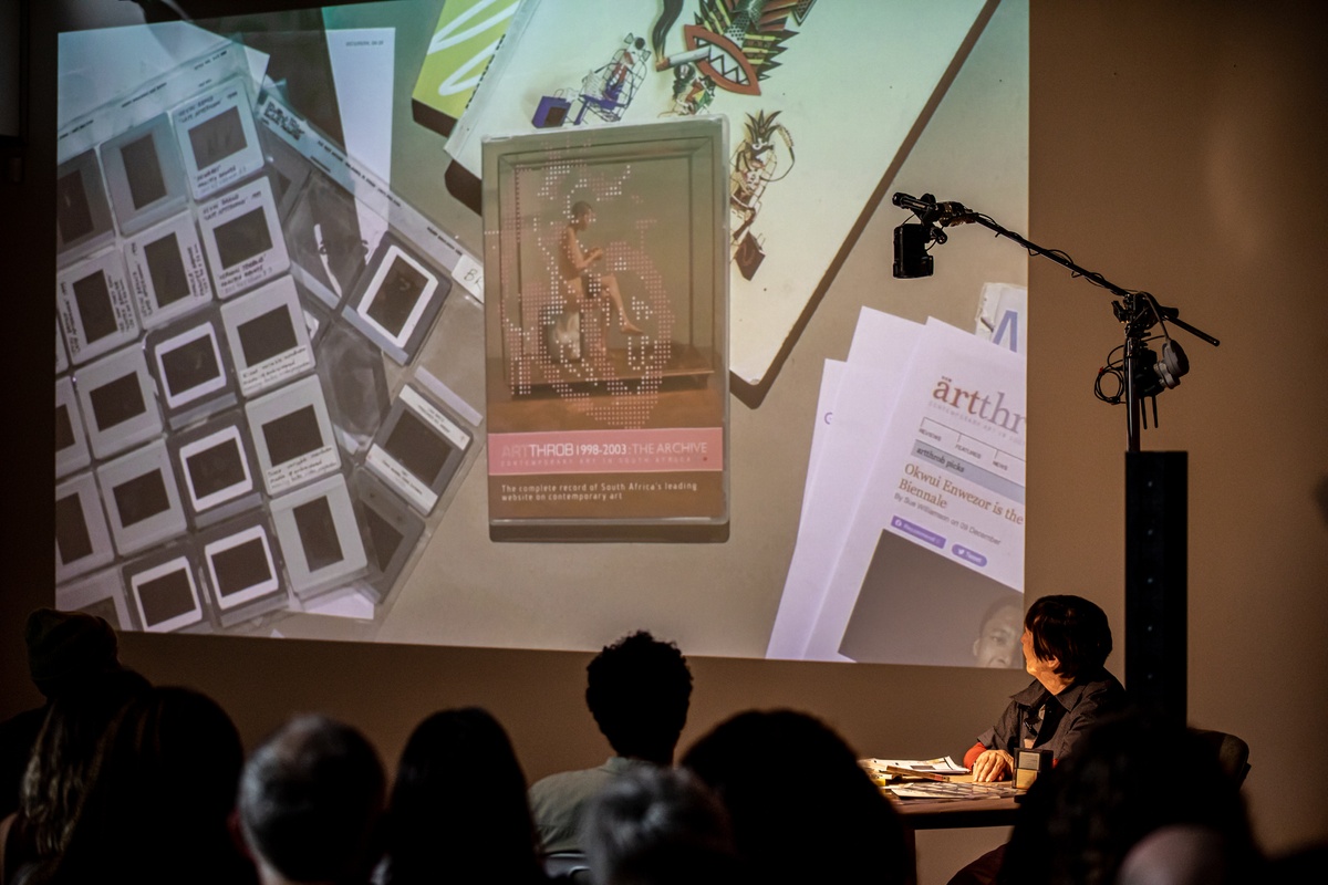 Event photograph from the book launch of 'ArtThrob: 25 Years of Art Writing in South Africa' in A4 Arts Foundation. At the front, attendees are seated. On the right, artist Sue Williamson is seated at the speaker's table. At the back, a livestream top-down view of the speaker's table is projected onto the wall.
