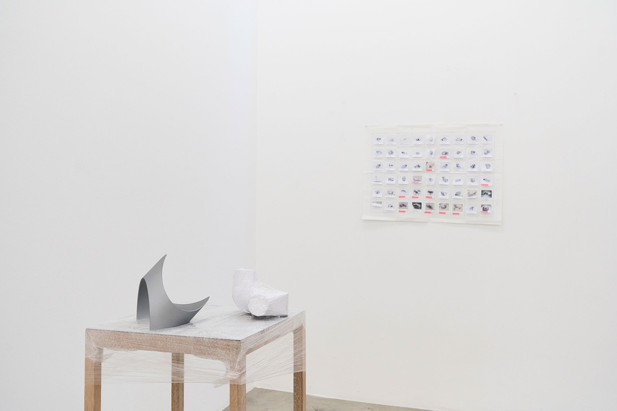 Process photograph from the 2nd rendition of ‘Parallel Play’ in A4’s Gallery. On the left, a display table wrapped in bubblewrap hosts two sculptural objects. On the right, small photographic snapshots with labels are arranged on a paper grid on a white wall.

