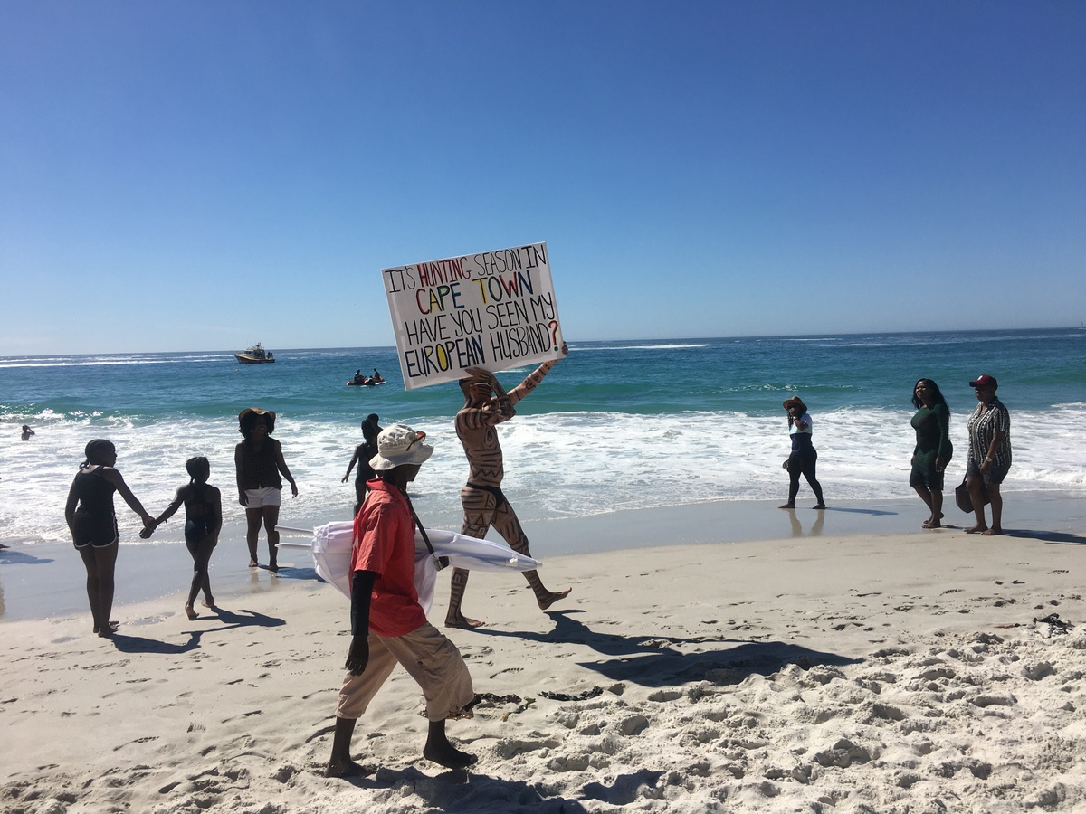 Event photograph from Kieron Jina’s ‘Catching Feelings, not Flights’ performance. The artist, their partially nude body covered in geometric designs, walks next to a Cape Town beach carrying a canvas that reads ‘It’s hunting season in Cape Town, have you seen my European husband?’ 
