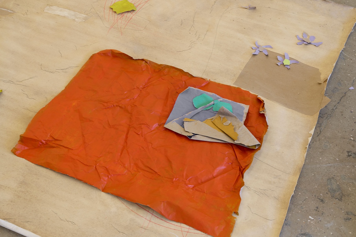 Process photograph of Asemahle Ntlonti’s working space during the 2nd rendition of ‘Parallel Play’ in A4’s Gallery. A large sheet of brown paper with drawing marks lines the floor, with a smaller sheet of paper painted orange lying on top of it.
