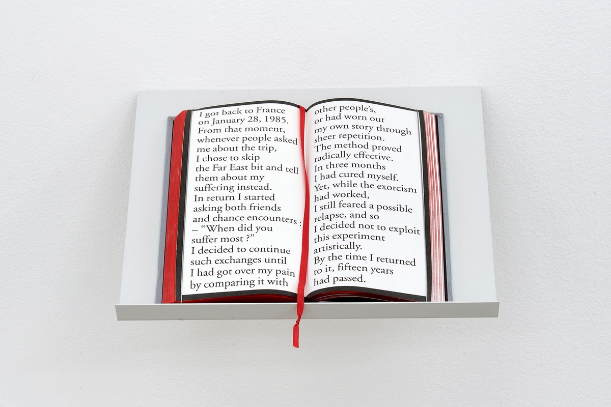 Installation photograph that shows a book from Sophie Calle’s installation ‘Exquisite Pain’ resting on a forward slanting wall-mounted shelf.
