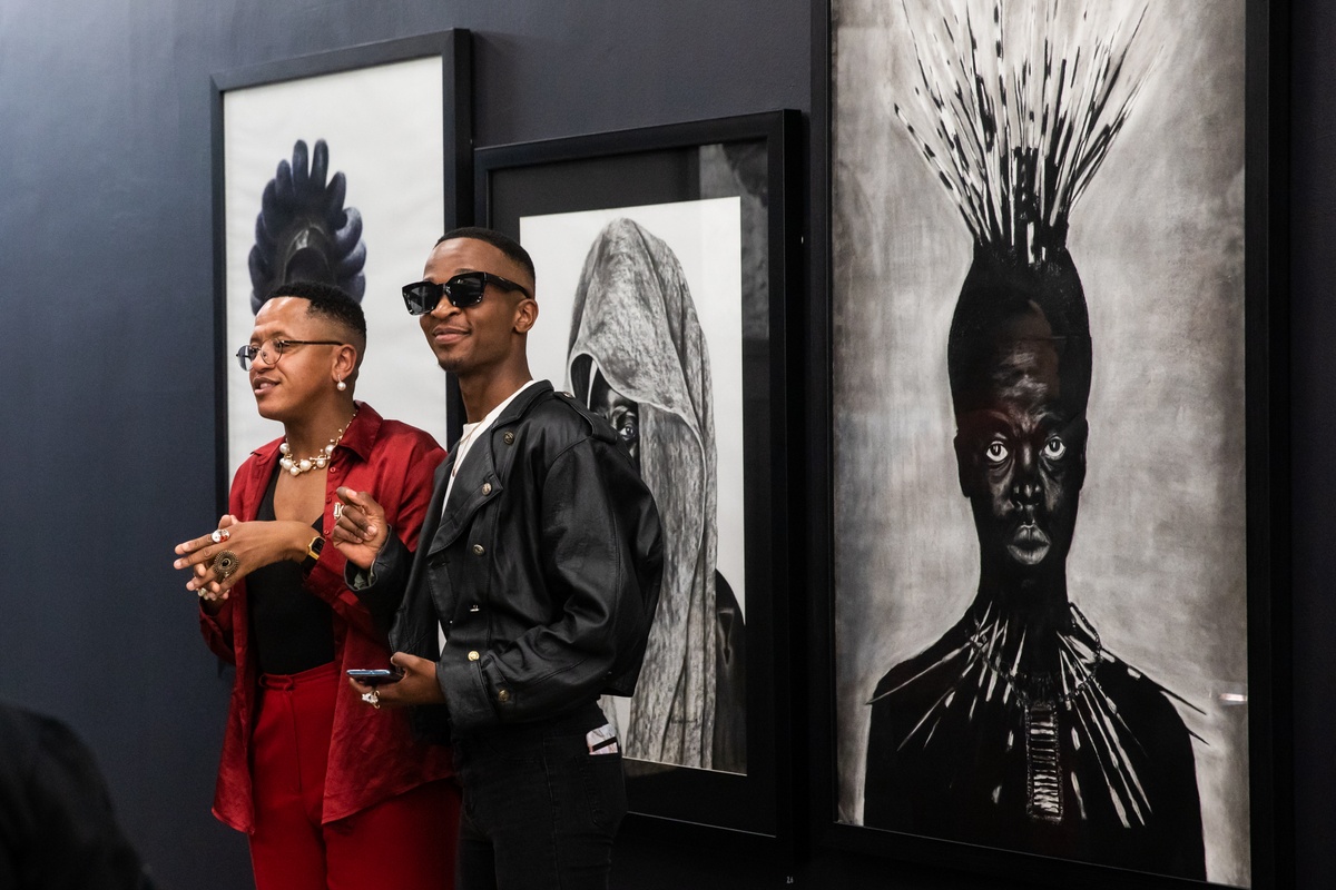 Event photograph from the opening of the “Ikhono LaseNatali” exhibition in A4’s Gallery. At the back, Lindani Nyandeni’s charcoal and ink drawing “Somnyama Ngonyama, ‘Ntozakhe II’,” Nhlanhla Chonco’s charcoal drawing “Somnyama Ngonyama, ‘Khwezi’” and Bongani Luthuli’s “Somnyama Ngonyama ‘Bester I’” are mounted on the black gallery wall. At the front, two attendees for the event.
