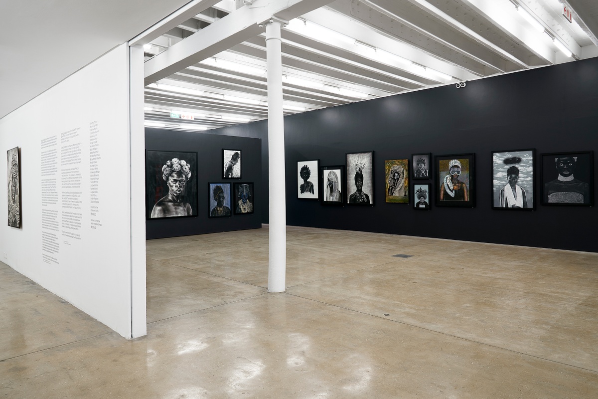 Installation photograph from the “Ikhono LaseNatali” exhibition in A4’s Gallery. On the left, an exhibition statement in vinyl is pasted onto a white gallery wall. On the right, various artworks line the black gallery walls.
