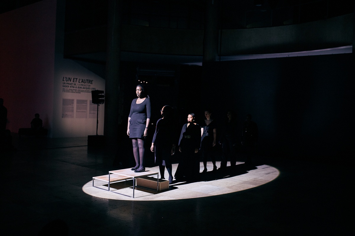 Event photograph from the offsite performance of ‘Elegy’ by Gabrielle Goliath at the D!STURB festival in Paris. A lineup of performers dressed in black stretches to a wooden podium with a performer standing on it on the left.
