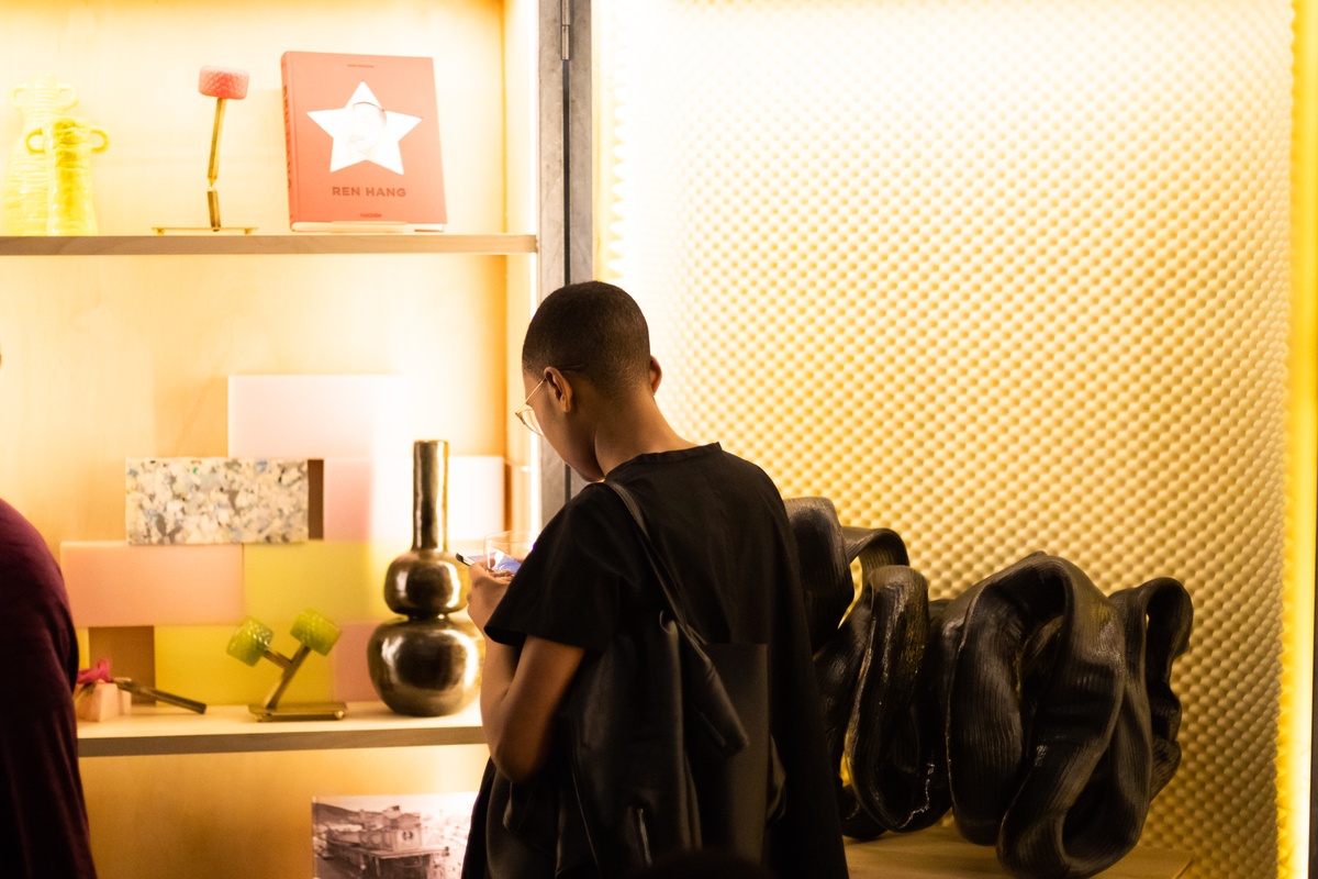 Event photograph from the ‘Hello proto~’ launch of A4’s Proto~ museum shop. In the middle, an attendee stands in front of a cabinet lined with various artworks and ephemera. At the top, Taschen’s Ren Hang photo-book sits on the shelf.
