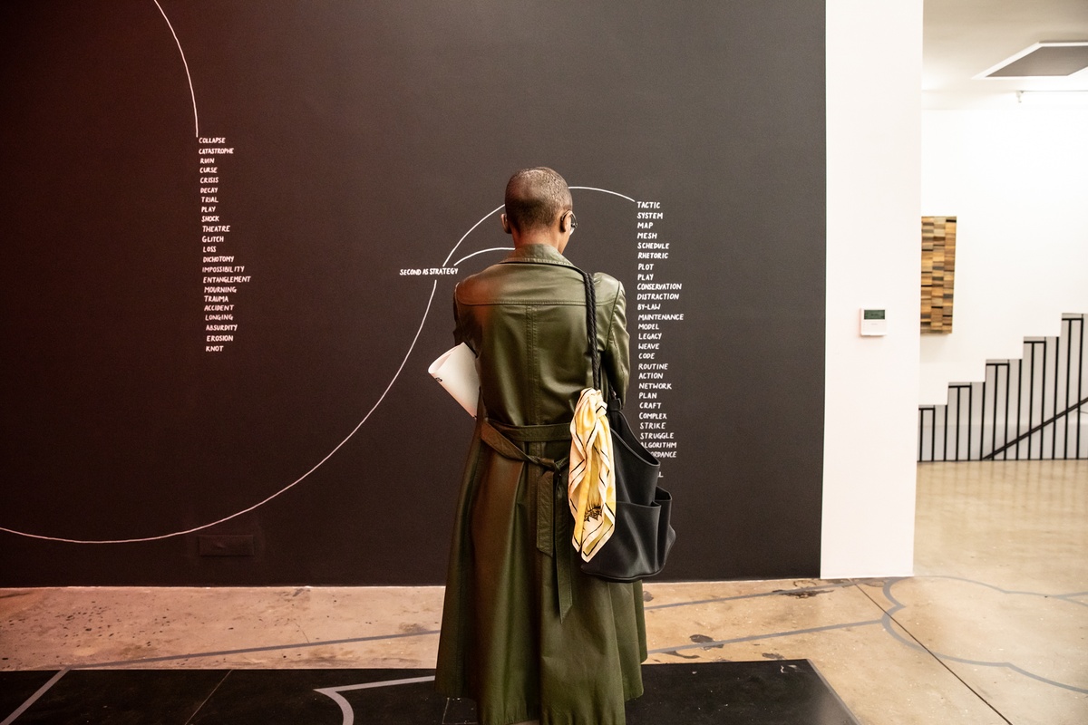 Event photograph from opening of the Customs exhibition in A4’s Gallery that shows an attendee standing in front of Nolan Oswald Dennis’ wall installation ‘Options’ on a black gallery wall.
