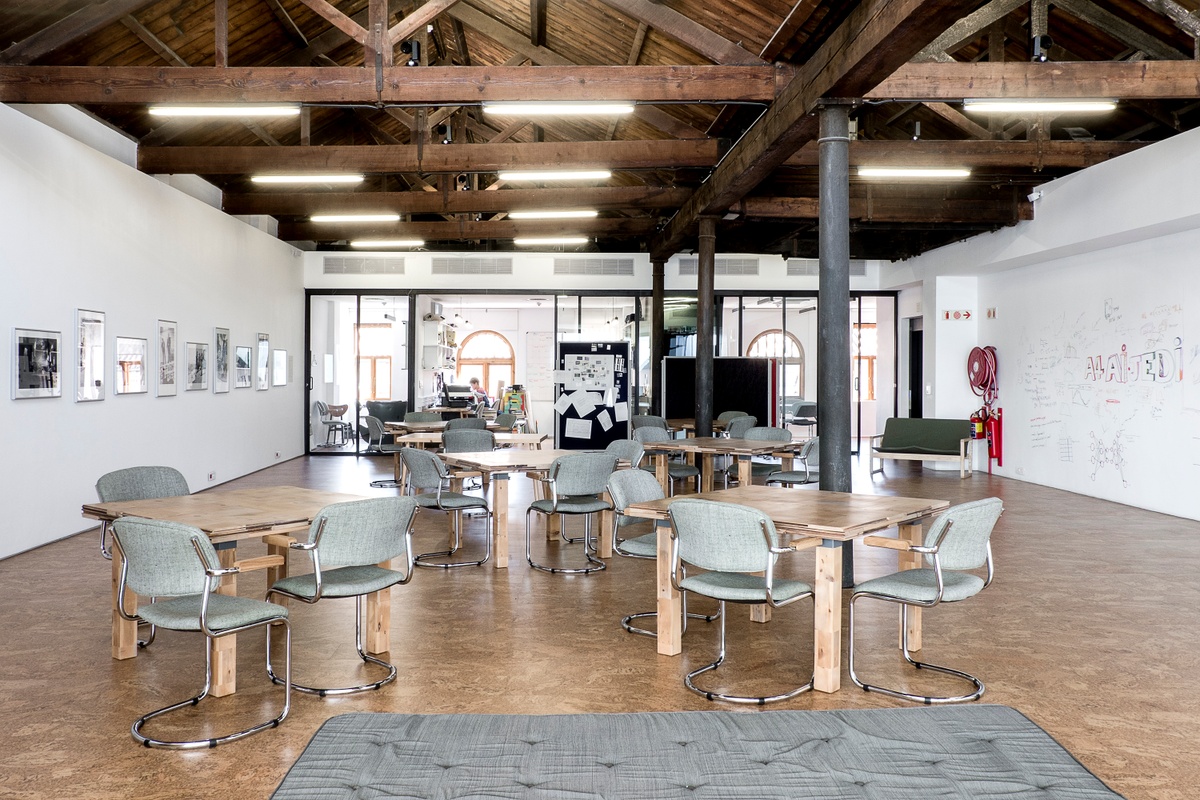 Installation photograph from the launch of Omar Badsha’s book ‘Seedtimes’ on A4’s top floor. In the middle, wooden tables with seating is arranged along the room. On the left, a row of framed monochrome photographs line the white wall. On the right, drawings line the wall.
