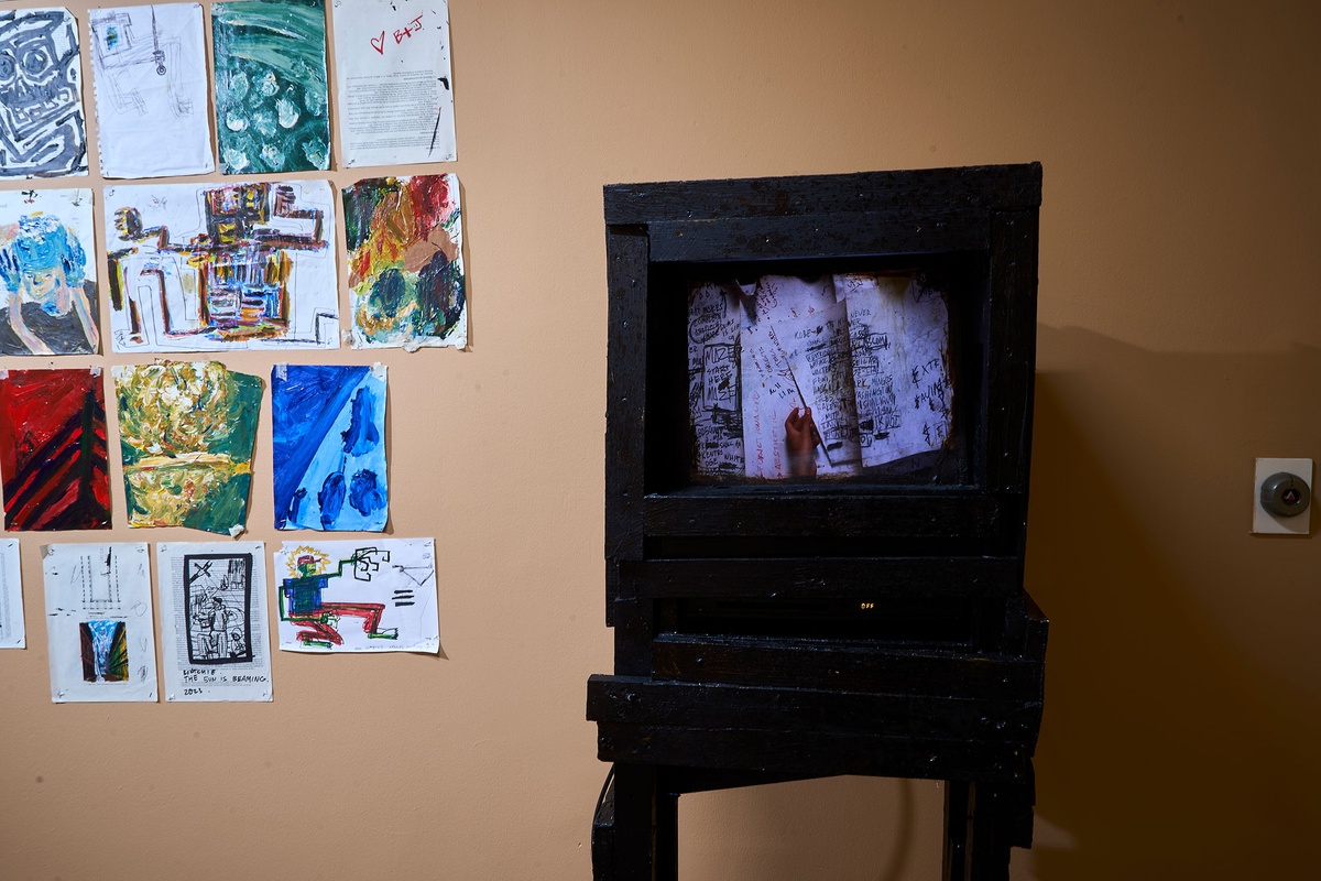 Installation photograph from Phillip Newman's exhibition in A4 Arts Foundation's Goods space. On the right, a video plays on a screen mounted in a wooden stand. On the left, drawings on paper are mounted on the wall.
