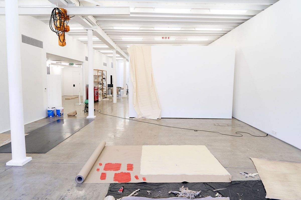 Process photograph of Asemahle Ntlonti’s working space during the 2nd rendition of ‘Parallel Play’ in A4’s Gallery. In the middle, sheets of brown paper and black plastic line the gallery floor. At the back, a sheet of white fabric hangs from a white movable wall.
