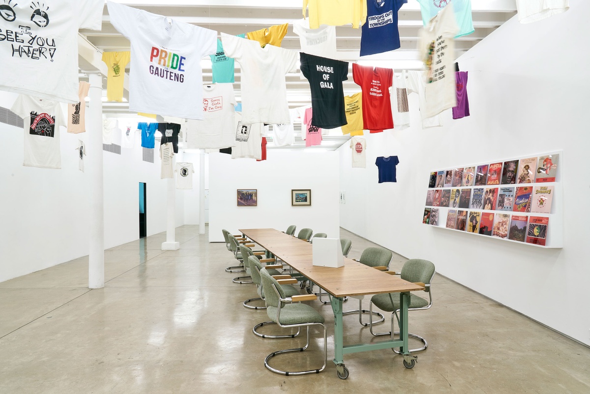 Installation photograph of the Common exhibition. On the right, vinyl record covers from ‘The Library of Things We Forgot to Remember’ by Kudzanai Chiurai are arranged on a slanted white shelf. In the middle is a long wooden table with an interactive box by Mitchell Messina. Above, T-shirts with slogans from the GALA Queer Archive and SAHA suspended on criss-crossing lines.
