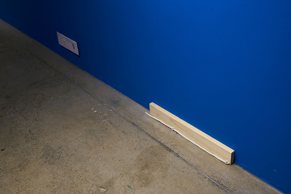 Installation photograph from Guy Simpson's Passage exhibition in A4 Arts Foundation's Goods space shows an unpainted wood and electric wire sculptural detail at the base of the blue wall.
