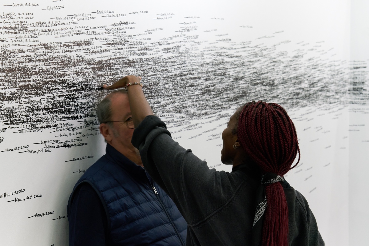 Event photograph of Roman Ondák’s performance piece ‘Measuring the Universe’ at A4’s booth at the 2020 Cape Town Art Fair. At the back, a white wall is covered with overlapping marks that indicate participant’s height, names and the date of their participation in black felt pen. At the front, an A4 facilitator measures the height of a fair attendee.
