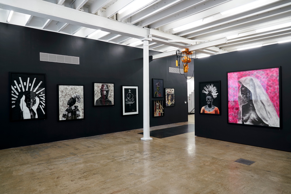 Installation photograph from the “Ikhono LaseNatali” exhibition in A4’s Gallery. On the right, Major Ndlovu’s acrylic painting “Somnyama Ngonyama, ‘Ndivile II’” and Mlamuli Sboniso Mkwanazi’s charcoal drawing “Somnyama Ngonyama” are mounted on a black movable gallery wall. On the left, various artworks line the black gallery wall.
