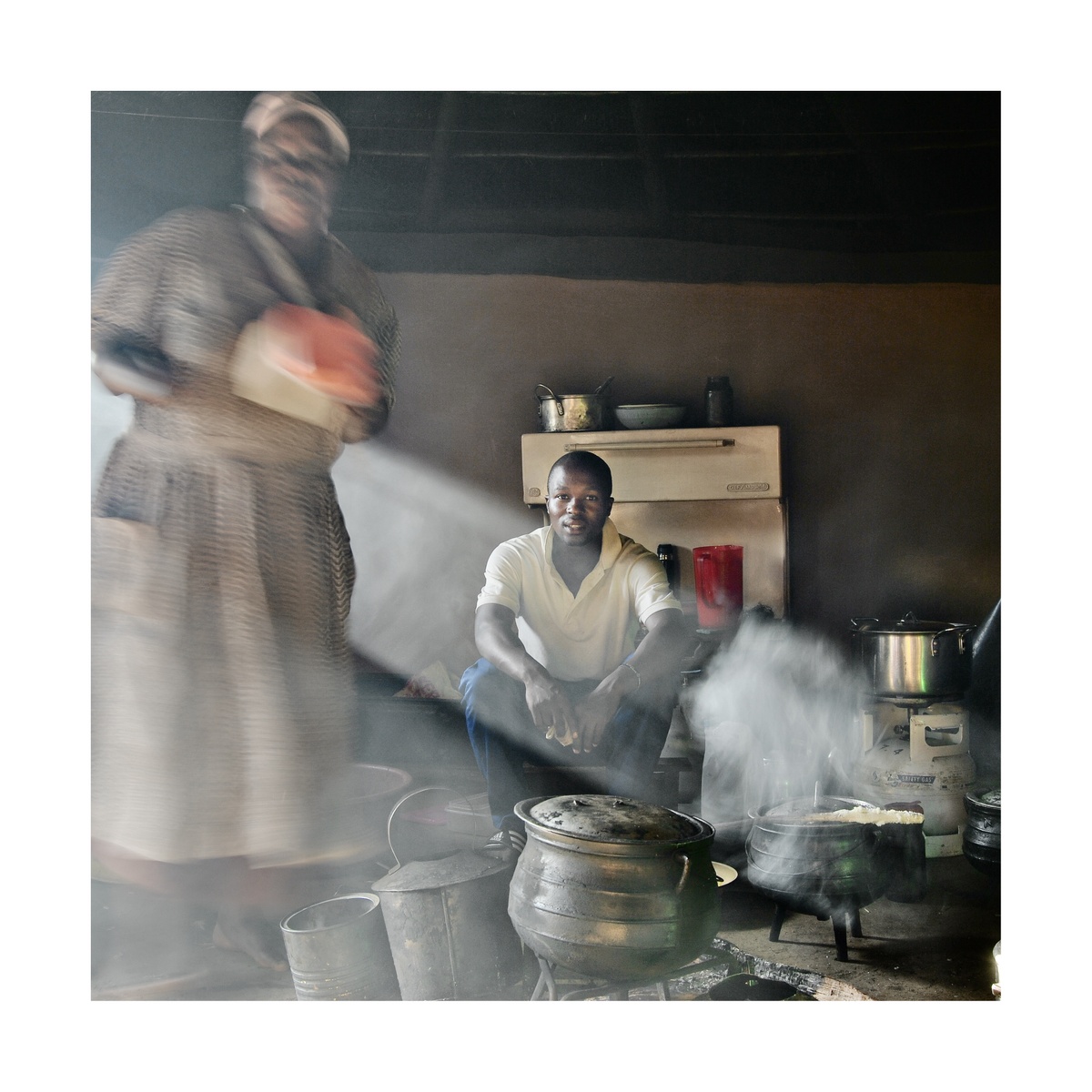 A photograph from Jabulani Dhlamini's residency on A4's top floor shows an indoor cooking area. At the back, one individual is seated. At the front, another individual stands tending various pots.
