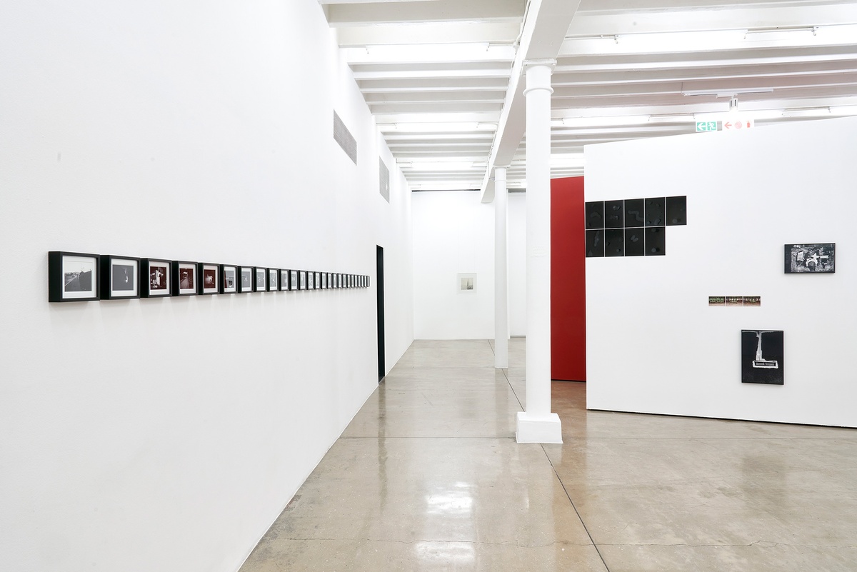 Installation photograph of the You to Me, Me to You exhibition. On the left, Bas Jan Ader’s 26 framed monochrome photographic prints ‘Study for In Search of the Miraculous (One Night in Los Angeles)’ are mounted on a white gallery wall in a straight line. On the right, Thembinkosi Hlatshwayo’s numerous photographic works are arranged on a white moveable gallery wall.
