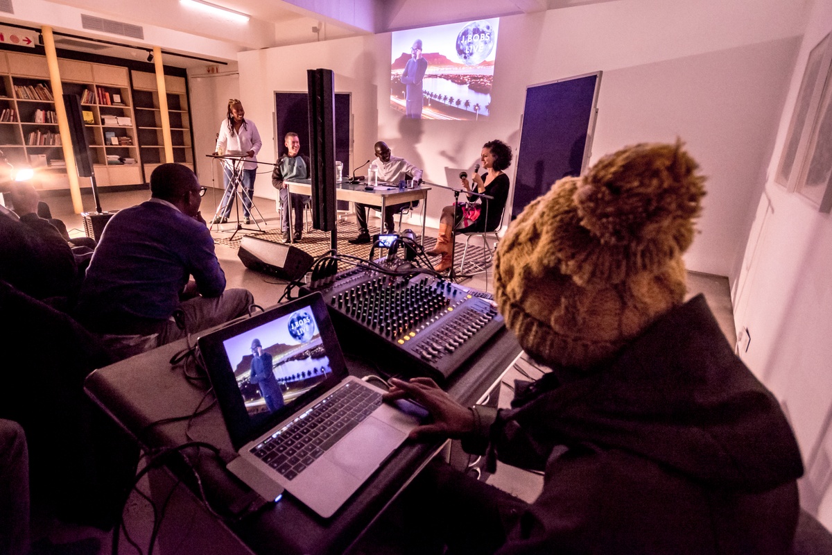 Event photograph from ‘A Game Show Double Bill’ by Kiri Pink Nob on A4’s ground floor. At the back, Jefferson Bobs Tshabalala is seated at a desk along with seated and standing participants, with a projection on the wall behind them. At the front, an individual operates the projection from a laptop, with a mixing board next to them.
