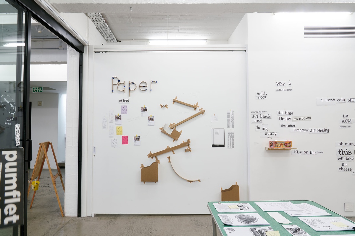 Installation photograph from the Papertrails exhibition in A4’s Reading Room. At the back, a cardboard Rube Goldberg machine is mounted to the wall, along with a cardboard relief of the word ‘Paper’.

