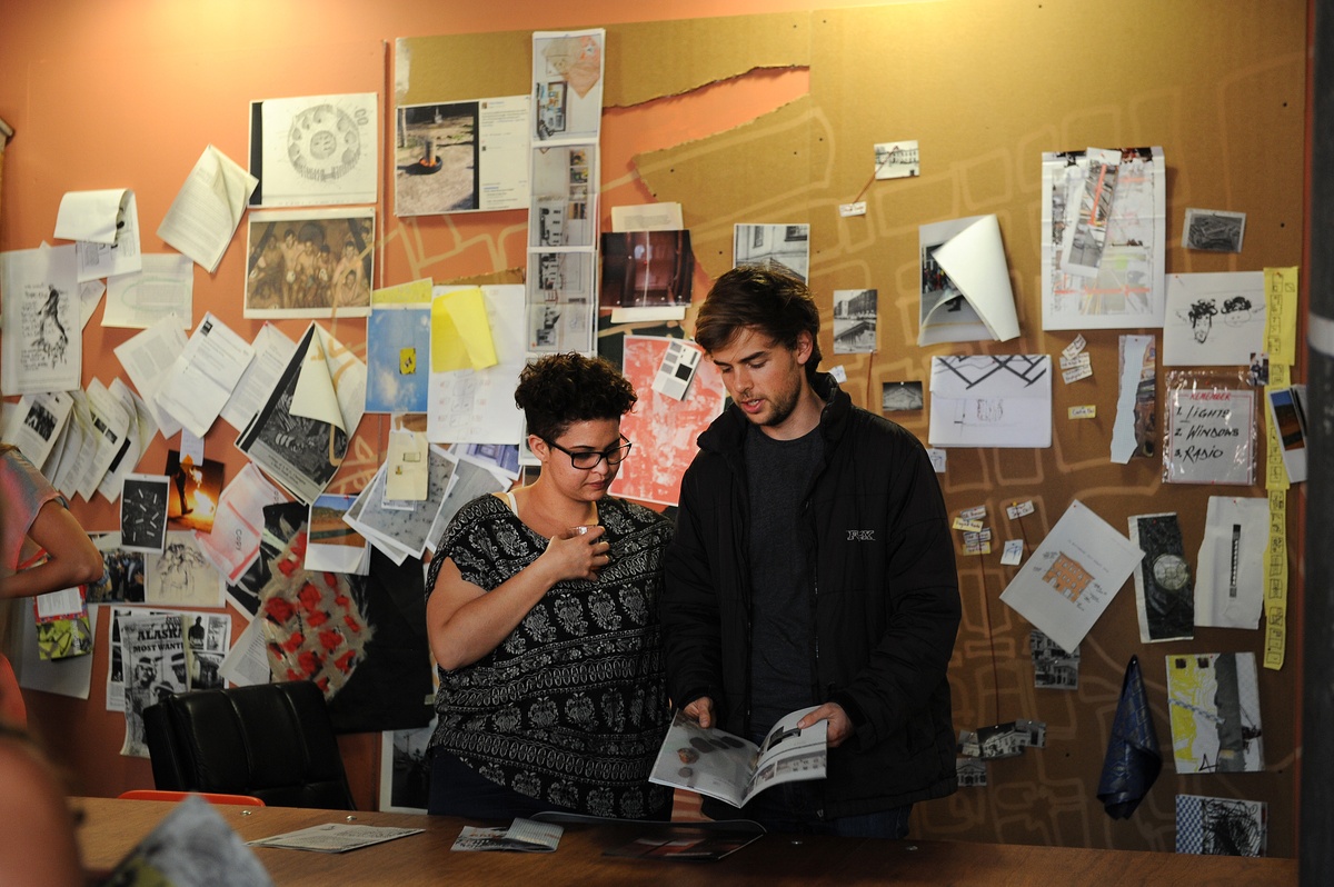 Event photograph from the offsite ‘Office Politics’ exchange. At the front, two attendees interact with copies of Tabogo George Mahashe and francis burger’s ‘Camera Obscura #3’ publication and Dorothee Kreutzfeldt’s ‘Landing Above’ publication a wooden table. At the back, the wall is lined with photocopies and drawings.
