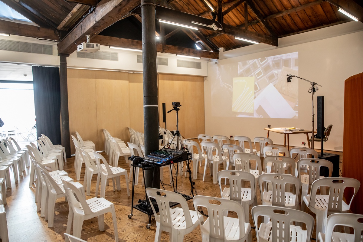 Event photograph from the book launch of 'ArtThrob: 25 Years of Art Writing in South Africa' in A4 Arts Foundation. At the front, rows of white chairs. At the back, a table for speakers with a livestream top-down view of it projected onto the wall on the left.
