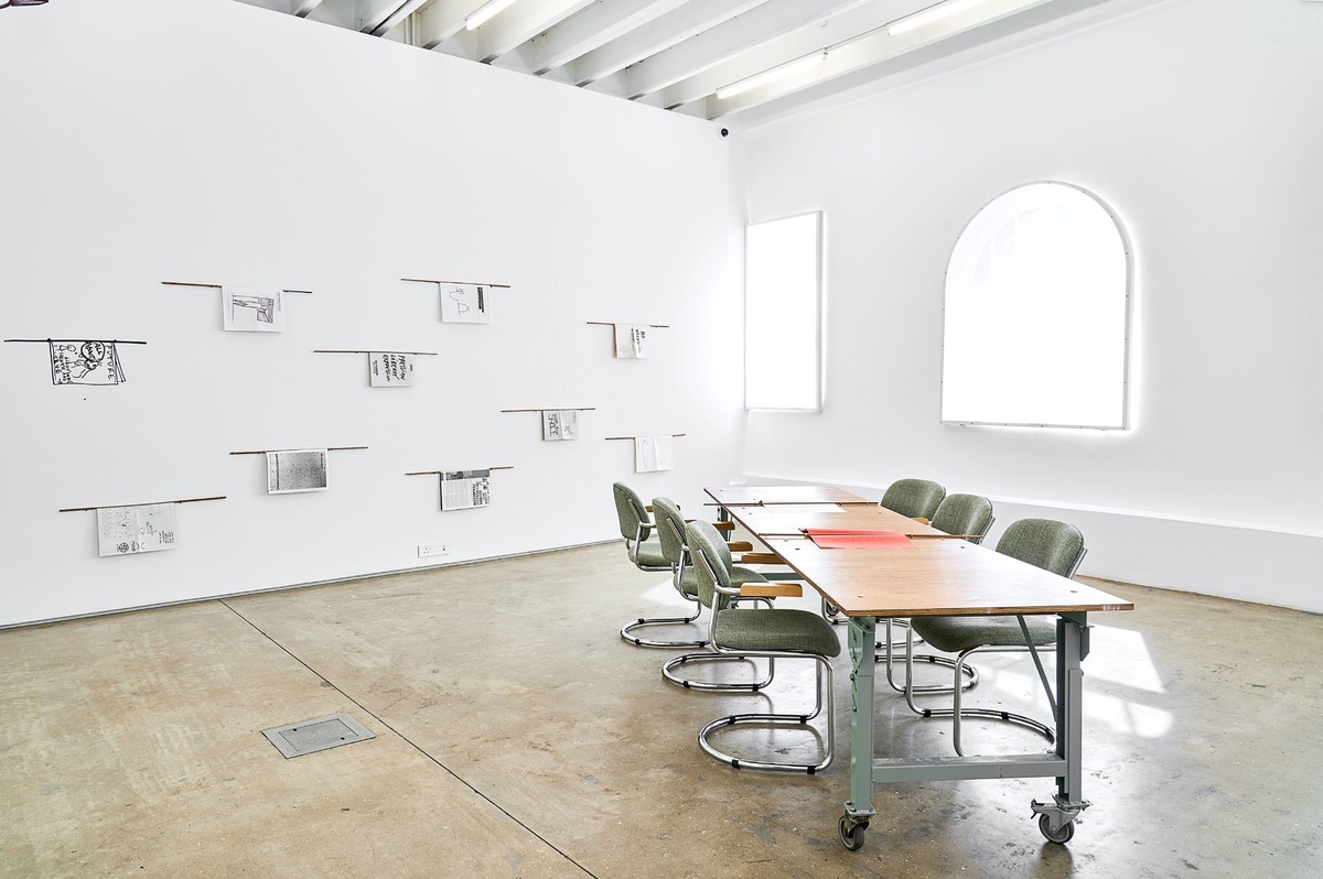 Installation photograph from Dan Perjovschi’s ‘The Black and White Cape Town Report’ exhibition in A4’s Gallery. On the left, the wall features a series of printed booklets suspended from wall mounted dowels. On the right, a long wooden table with seating.
